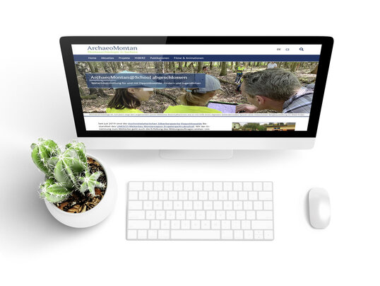 Bildschirm mit der ArchaeoMontan-Website, davor Tastatur, Maus und ein Kaktus in einem weißen Blumentopf