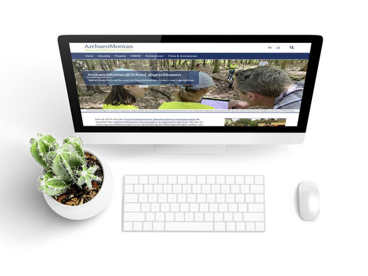 Bildschirm mit der ArchaeoMontan-Website, davor Tastatur, Maus und ein Kaktus in einem weißen Blumentopf